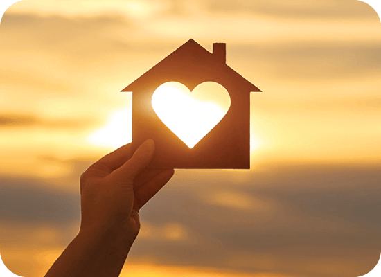 A hand holds a silhouette of a house with a heart cutout against a sunset background.